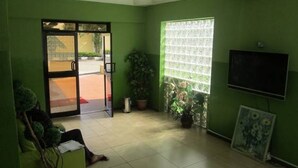 A room with green walls, a tiled floor, a door leading outside, a window with blinds, a potted plant, and a flat-screen TV mounted on the wall. - Lamonde Guest House (Owerri)