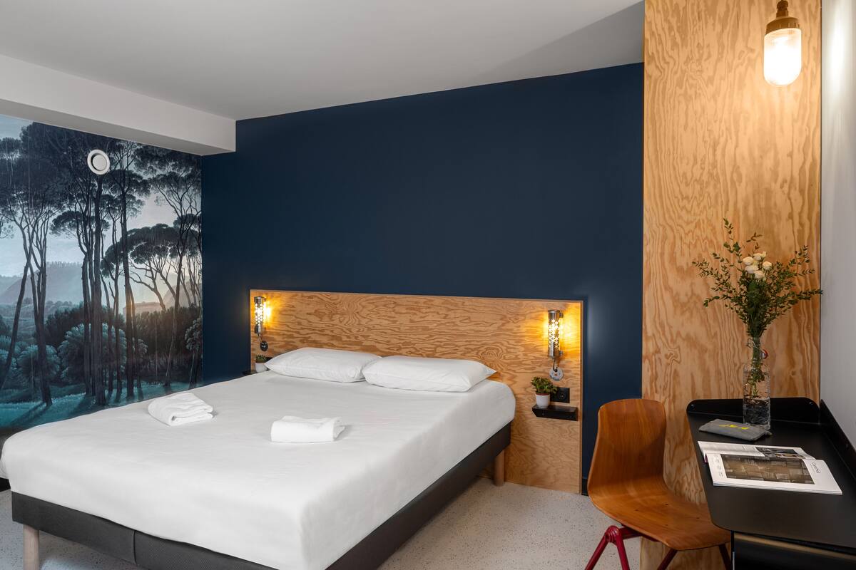 A hotel room with a large bed, a nightstand, a desk with a chair, and a wall-mounted lamp.