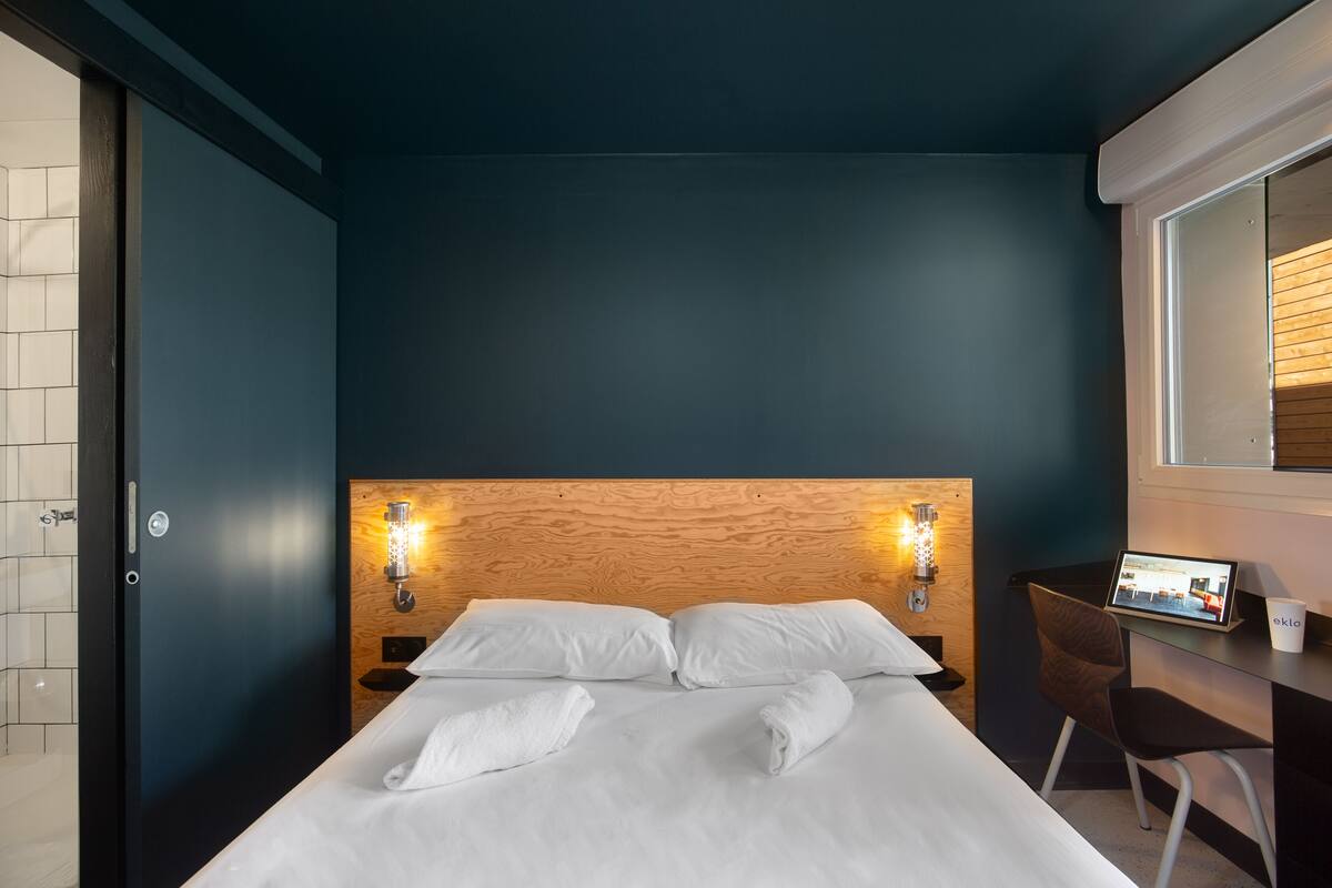 A hotel room with a bed, two bedside lamps, a desk with a computer, and a window with blinds.