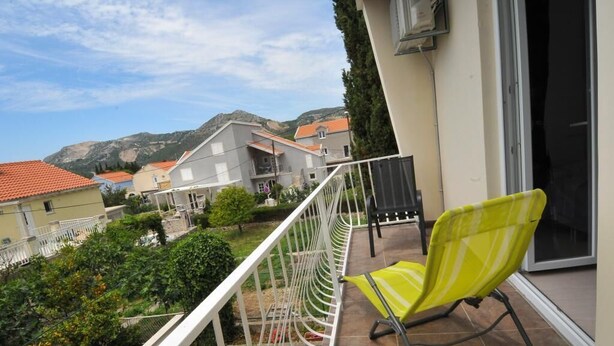 Un balcon avec une chaise jaune, une balustrade blanche et une vue sur des maisons et de la verdure en arrière-plan.