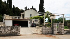Une entrée fermée par un portail et bordée d’un mur en pierre, un bâtiment blanc avec un garage, et une voiture garée à l’intérieur.