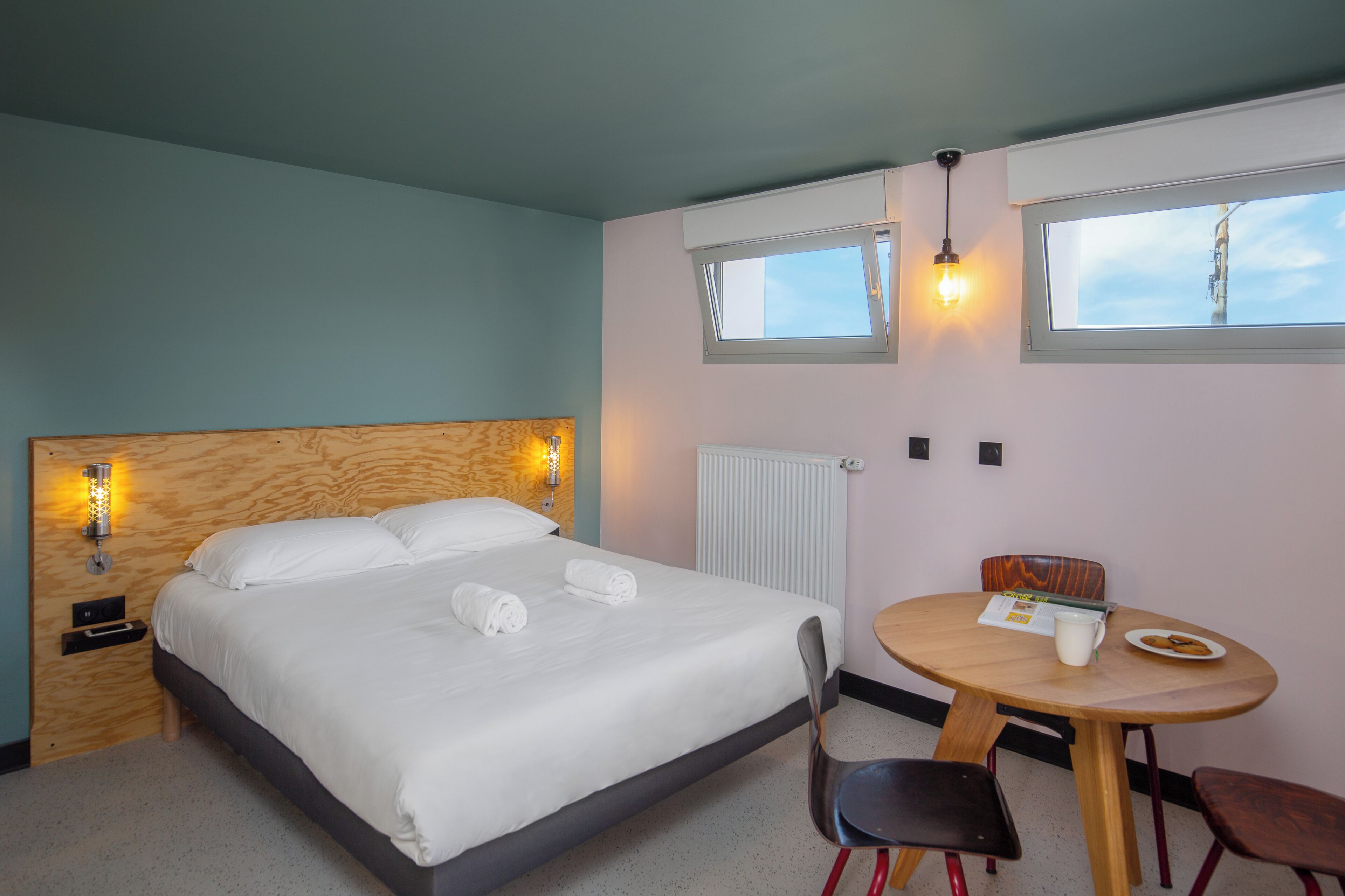 A hotel room with a bed, a table with a chair, a window, and a wall-mounted light fixture.