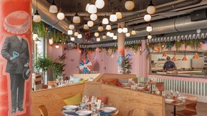 Un restaurant avec des tables rondes et des banquettes, un chef derrière le comptoir, et un mur coloré orné d’une roue qui tourne.