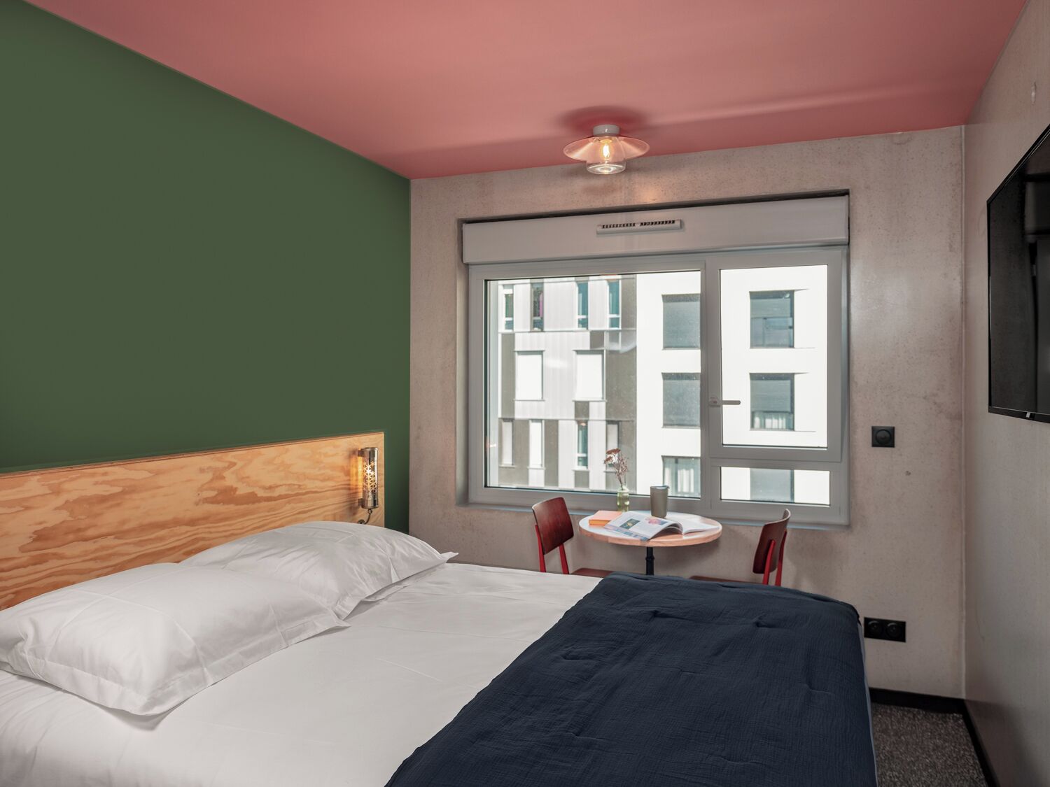 Une chambre d’hôtel avec un lit, une petite table et une fenêtre donnant sur des immeubles.