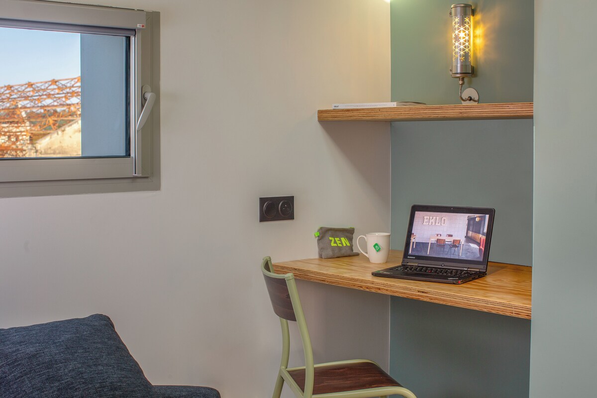 Un espacio de trabajo pequeño y ordenado con una computadora portátil, una taza y una bolsa tipo Zen sobre una estantería de madera, una silla y una ventana que da a una obra en construcción.