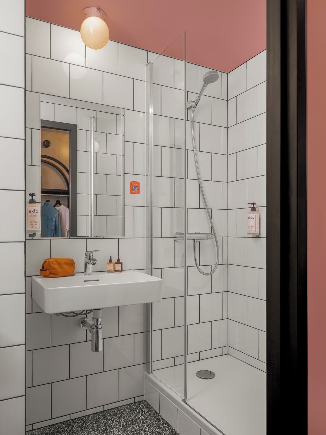 Een moderne badkamer met een douchecabine van glas, een witte wastafel met een spiegel erboven en toiletartikelen op de wastafel.