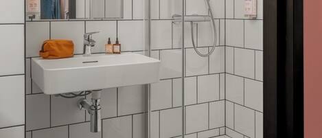 Een moderne badkamer met een douchecabine van glas, een witte wastafel met een spiegel erboven en toiletartikelen op de wastafel.