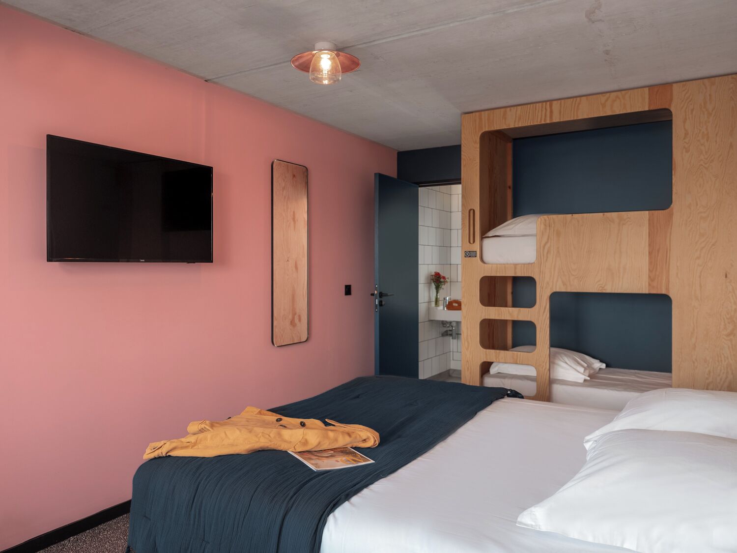 Habitación de hotel con pared rosa, televisión de pantalla plana, cama litera de madera y mesita de noche.