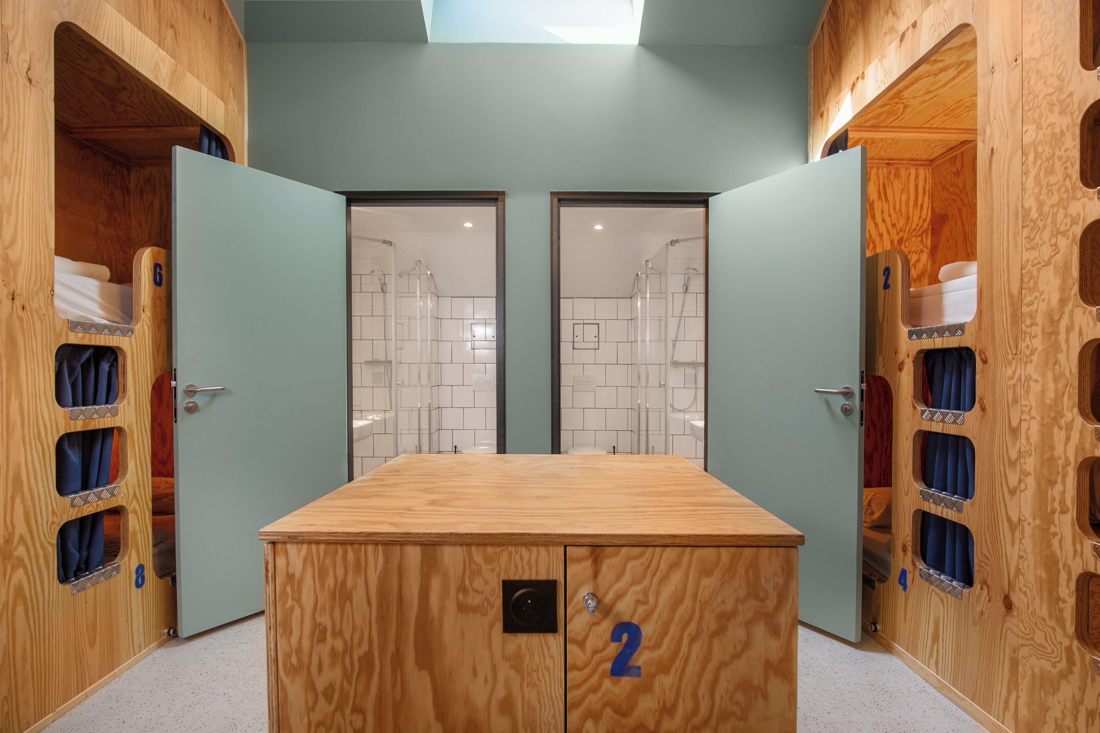A room with two wooden bunk beds, a central wooden table, and a shower area visible through a glass door.