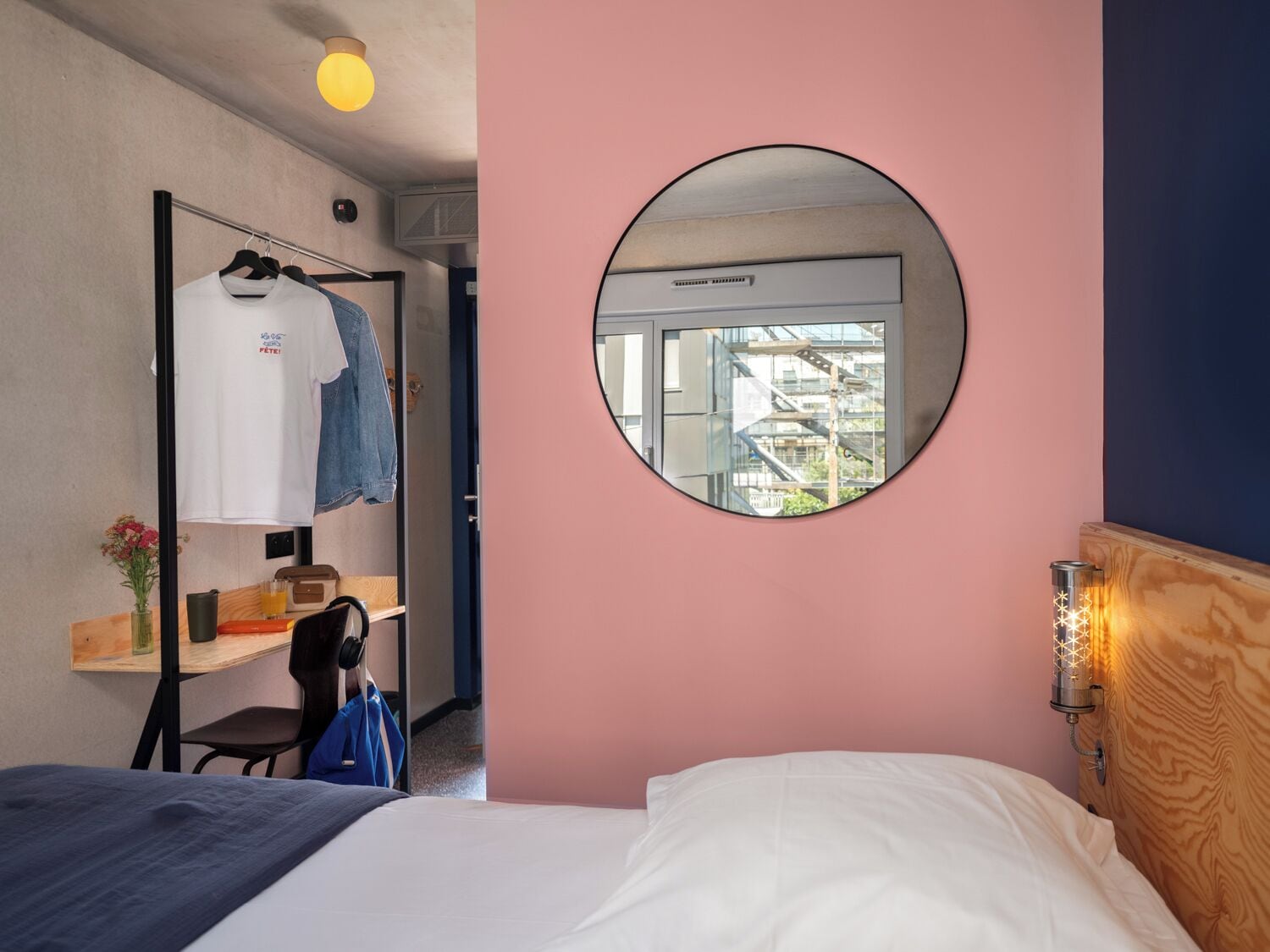 Una habitación de hotel con una cama, un escritorio con silla, un armario, un espejo redondo y una ventana con vista a un edificio.