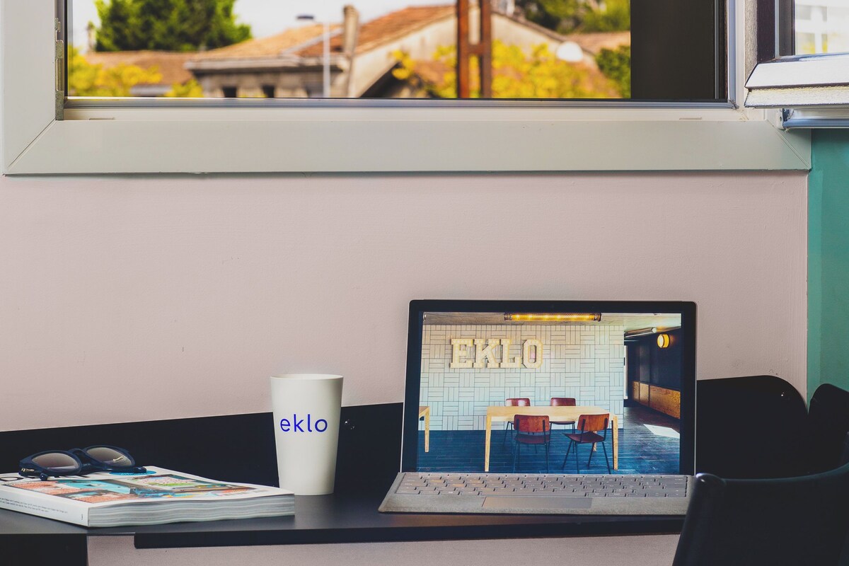 Un ordinateur portable est posé sur un bureau, et par la fenêtre, on peut voir un bâtiment et des arbres.