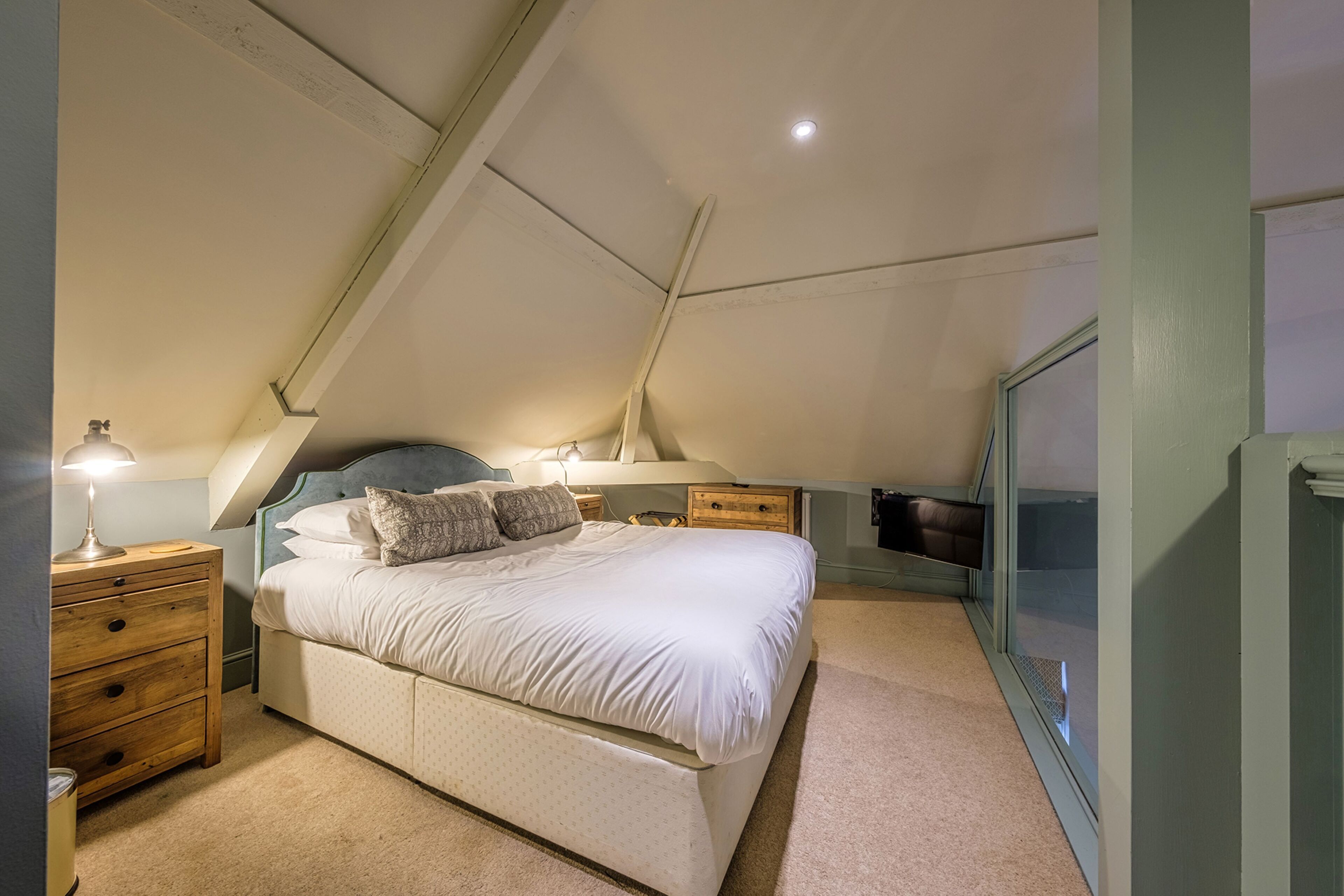 A bedroom with a sloped ceiling, a bed with white bedding, a bedside table with a lamp, and a dresser.