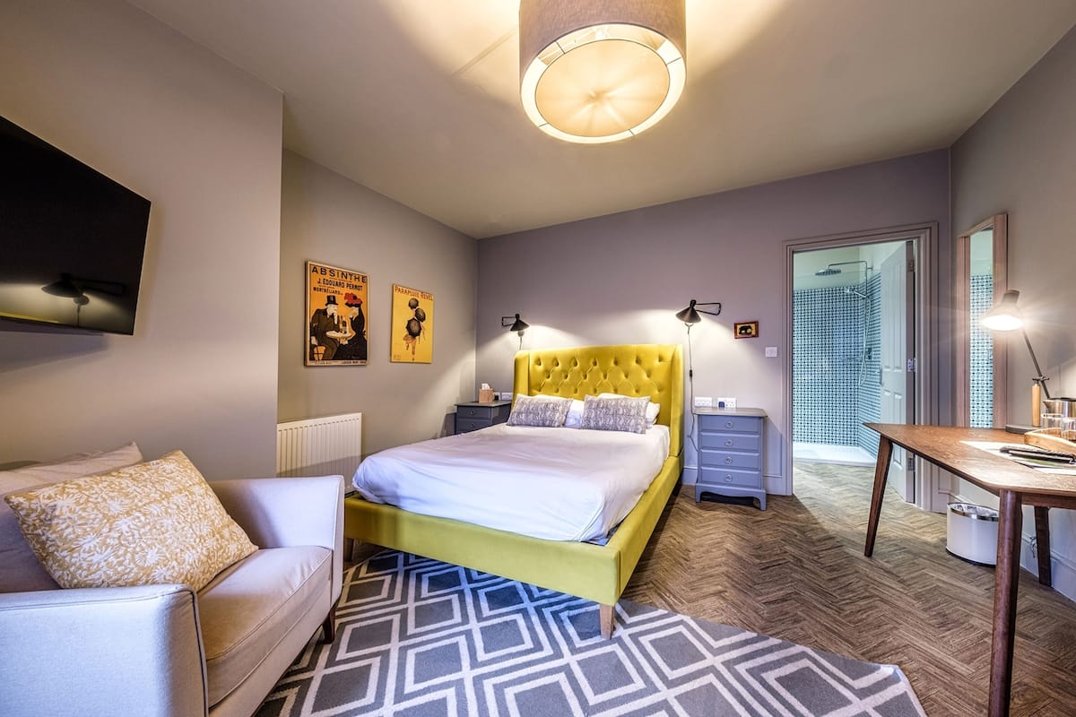 Un dormitorio con cama amarilla, sofá blanco, alfombra estampada y televisor montado en la pared.