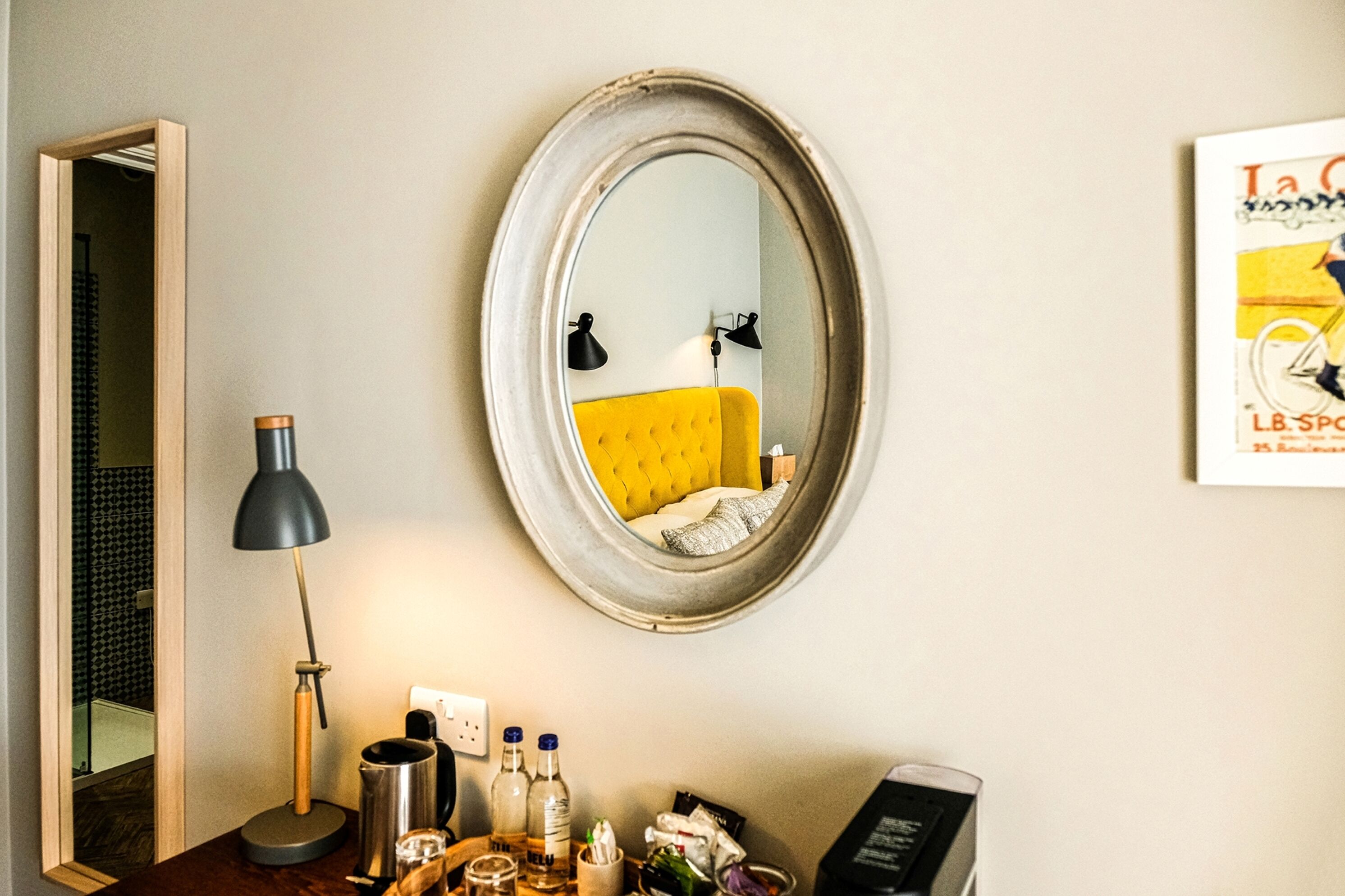 A room with a round mirror, a yellow sofa, a desk with a lamp, and a framed picture on the wall.
