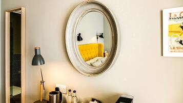 A room with a round mirror, a yellow sofa, a desk with a lamp, and a framed picture on the wall.