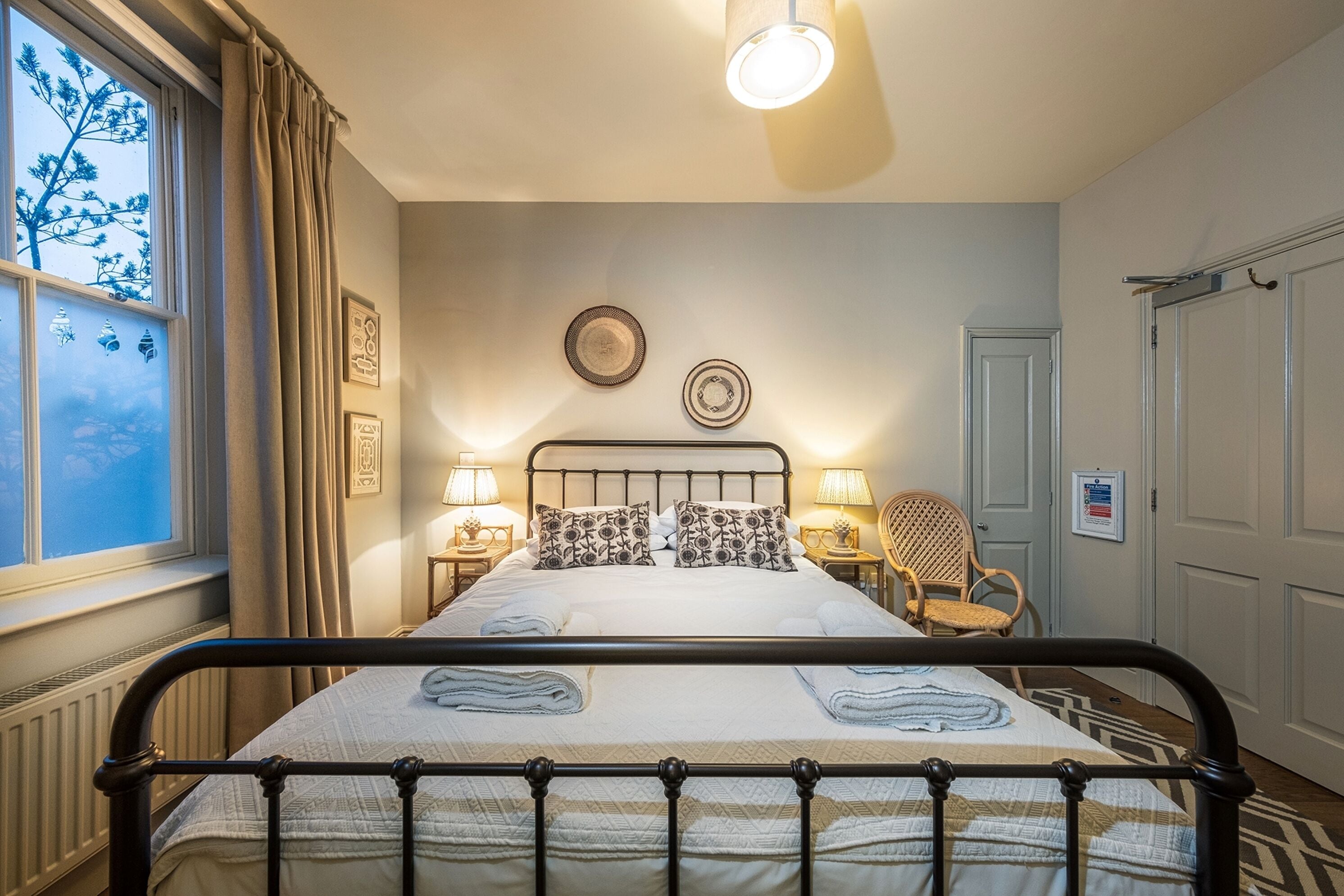 A neatly made bed with a headboard, two bedside lamps, a chair, and a window with curtains.