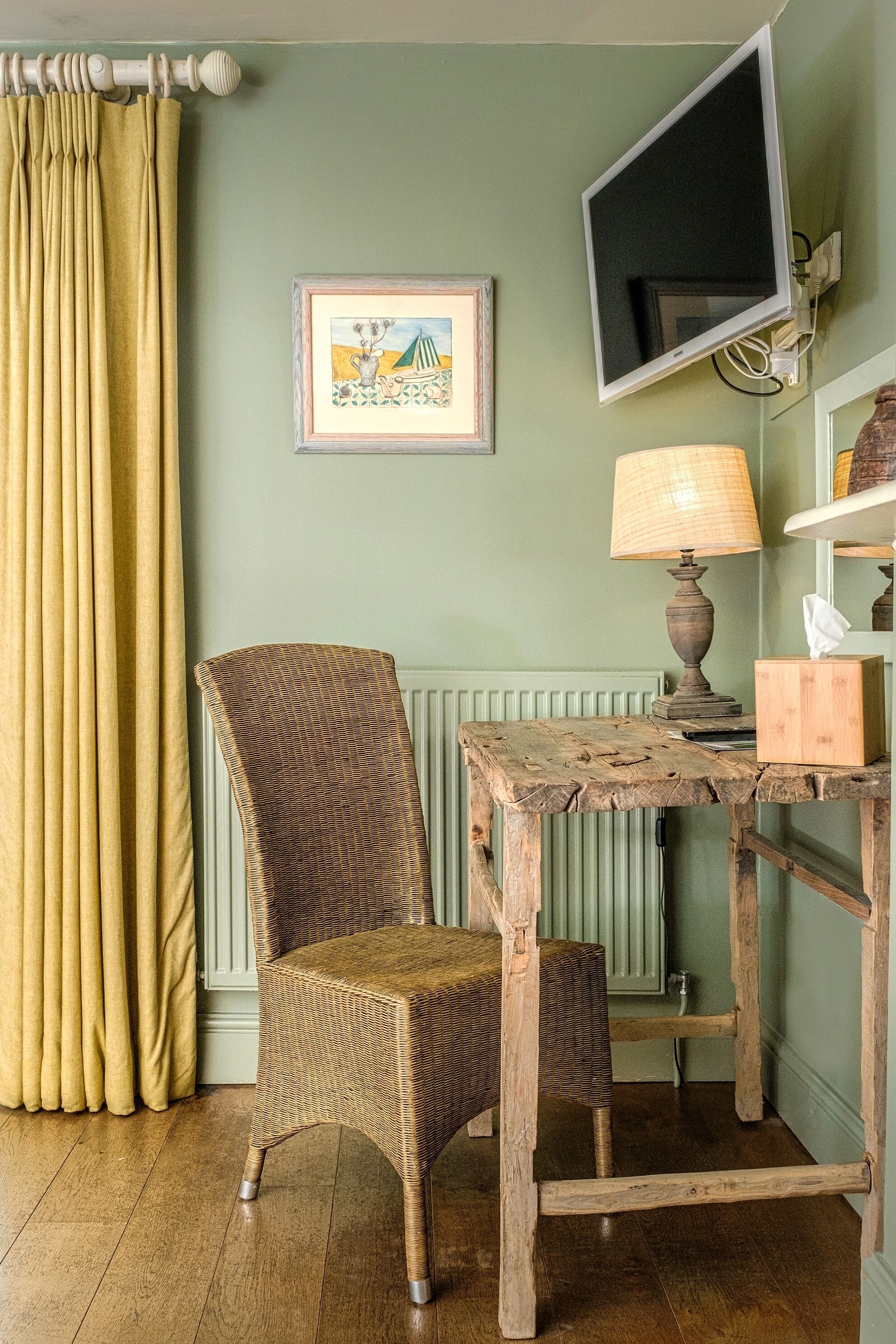 Una habitación con una silla de mimbre, una mesa de madera, una lámpara, un cuadro enmarcado y un televisor en la pared.