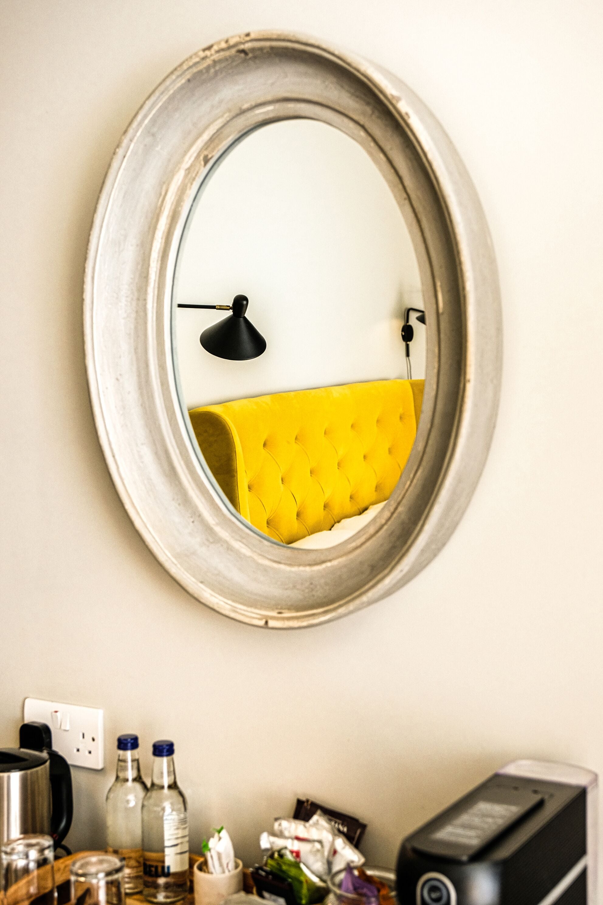 A round mirror with a white frame, a yellow upholstered bench, a black desk lamp, and a black printer on a desk.