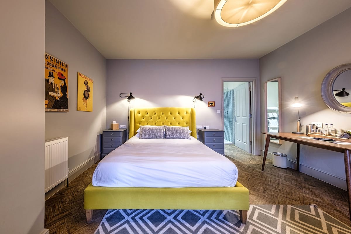 Un dormitorio moderno con un cabecero tapizado amarillo, una cama grande, mesitas de noche, un escritorio y un espejo.