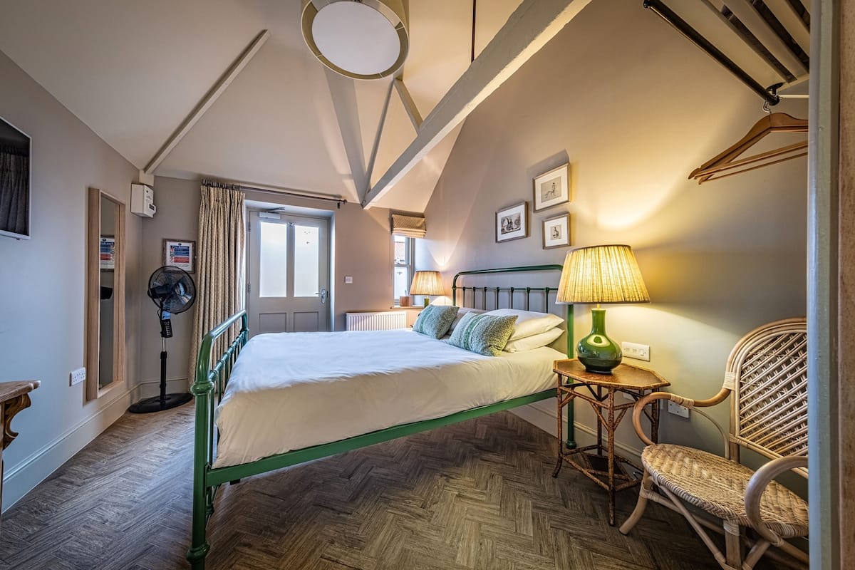 Un dormitorio con cama, una silla, una mesa con lámpara y un ventilador.