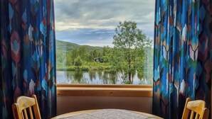 A room with a round wooden table, two wooden chairs, a window with a view of a lake and hills, and patterned curtains. - Vauldalen Fjellhotell (Roros)