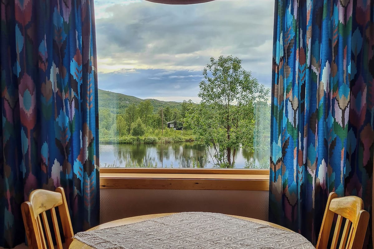 Une pièce avec une table ronde en bois, deux chaises en bois, une fenêtre donnant sur un lac et des collines, et des rideaux à motifs.