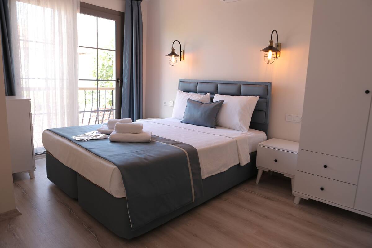 A neatly made bed with a grey and white duvet, two bedside tables, a wardrobe, and a window with curtains.