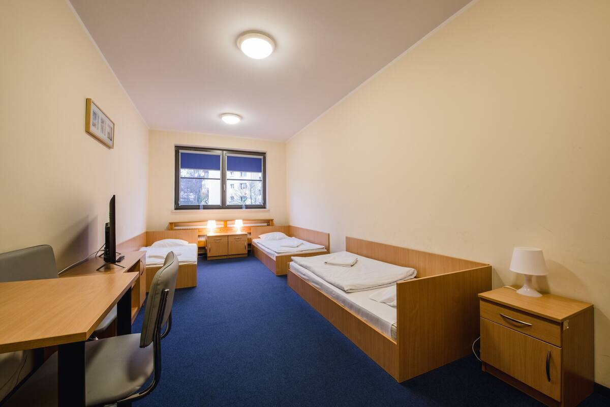 Habitación de hotel con dos camas individuales, un escritorio con silla, un mueble bajo y una ventana con vistas a edificios.