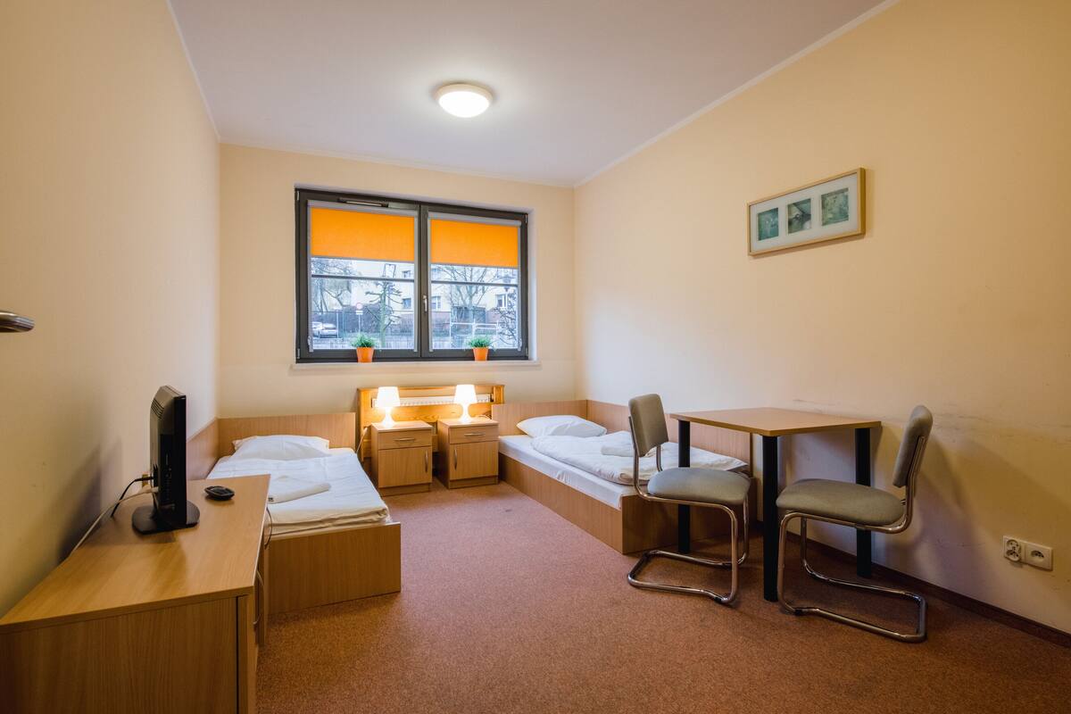 Una habitación pequeña y bien iluminada con una cama individual, un escritorio, una silla y una ventana con persianas anaranjadas.