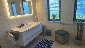 A modern bathroom with a white vanity, a large mirror with LED lighting, two windows with curtains, and a potted plant. - Vacation apartment Pier 7 - directly on the lake! (Bitterfeld-Wolfen)