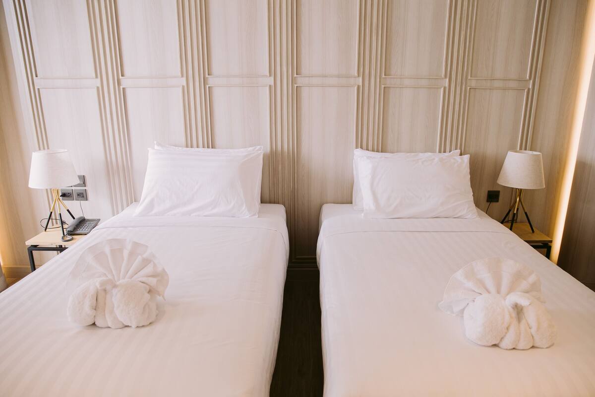 Habitación de hotel con dos camas, cada una con ropa blanca y almohadas, y una toalla sobre cada cama. Hay lámparas de noche y un teléfono en la mesita de noche.