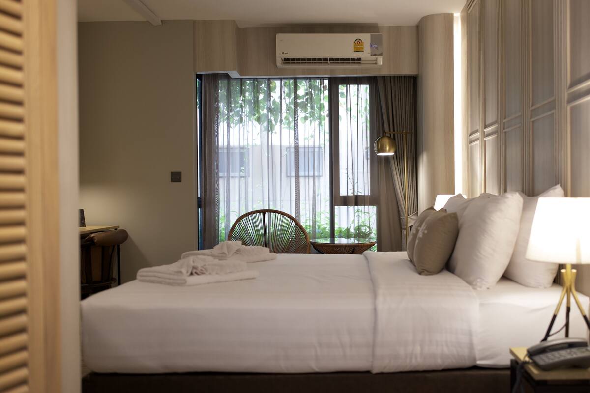 Habitación de hotel con una cama grande, una mesita de noche con lámpara, una silla y una ventana con cortinas.