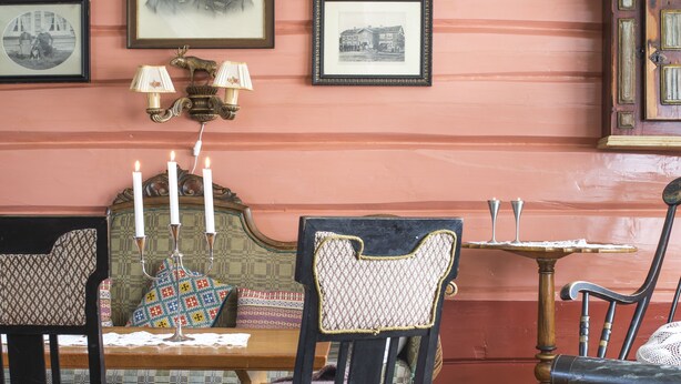 Une salle à manger avec une table en bois, deux chaises, un banc et un fauteuil à bascule. Des tableaux encadrés ornent le mur et un lustre est suspendu au-dessus de la table.