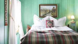 Un lit avec du linge de lit blanc, une couverture à carreaux et un coussin décoratif. Un tableau représentant un paysage est accroché au mur au-dessus du lit.