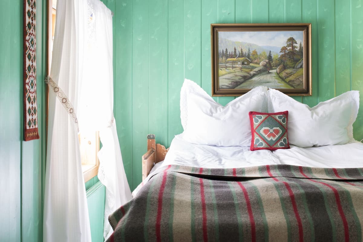 Una cama con sábanas blancas, un edredón a cuadros y una almohada decorativa. Un cuadro de un paisaje cuelga de la pared sobre la cama.