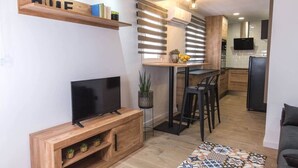 Ett modernt vardagsrum med en TV, ett träinrett mediaskåp och en köksdel med ett matbord och stolar.