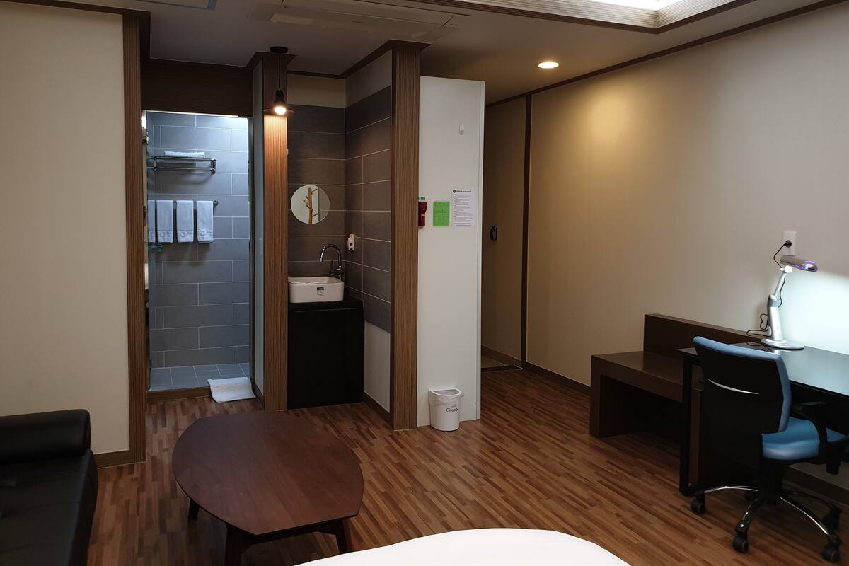 Una habitación de hotel con piso de madera, un escritorio con una silla, un lavamanos pequeño y un espejo.