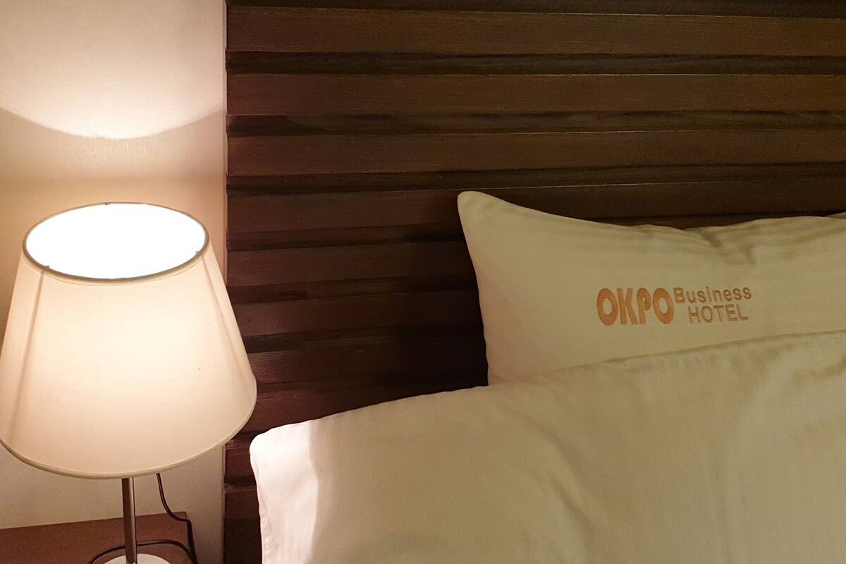 Habitación de hotel con cabecera de madera, almohada blanca con el bordado 'OKPO Business Hotel' y una lámpara de noche.