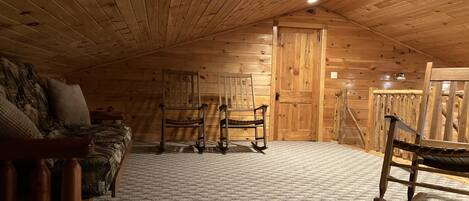 A cozy attic bedroom with wooden walls and ceiling, a bed with patterned bedding, a rocking chair, and a small table.