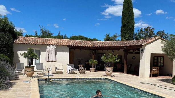 A swimming pool with a pink flamingo float, a person swimming, and a covered patio area with chairs and umbrellas. - Mas provençal en pierre  (UZES)