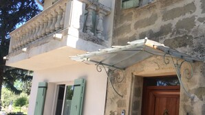 A two-story building with a balcony, a wooden door, and a table with a chair outside. - Mas provençal en pierre  (UZES)