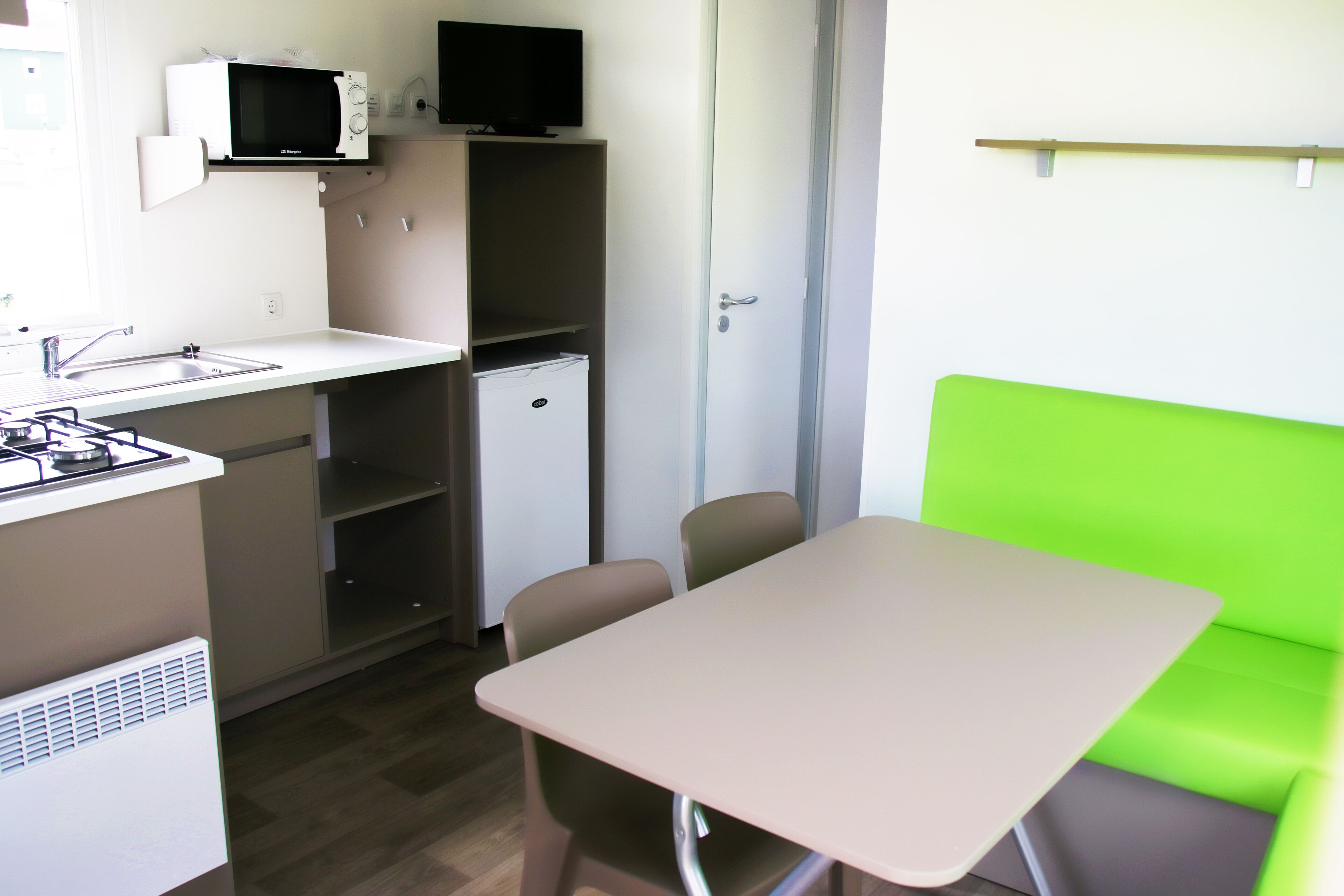 Cocina compacta equipada con microondas, lavamanos y un refrigerador pequeño. Hay una mesa con sillas y un banco con cojín verde.