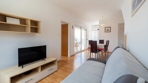 A modern living room with a flat-screen TV on a wooden stand, a grey sofa, and a dining area with a table and chairs. - Apartments Fran (Vela Luka)