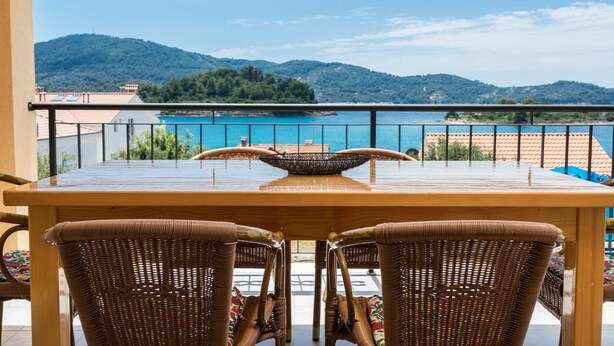 A wooden table with wicker chairs on a balcony overlooking a turquoise sea and green hills. - Apartments Fran (Vela Luka)