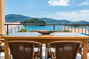 A wooden table with wicker chairs on a balcony overlooking a turquoise sea and green hills. - Apartments Fran (Vela Luka)