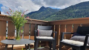 Um deck de madeira com duas cadeiras e uma mesa, um vaso com flores e vista para as montanhas.