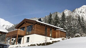 Uma cabana de madeira com varanda, cercada por árvores e montanhas cobertas de neve.