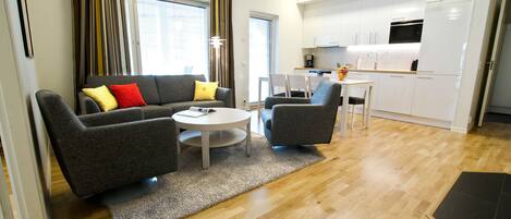 A modern living room with a grey sofa, two armchairs, and a small round table. A kitchen area with white cabinets and a microwave is visible in the background.