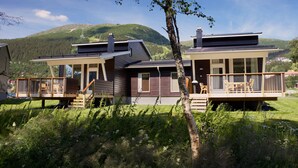 A modern two-story house with a deck and large windows, surrounded by a lush green landscape and hills in the background.