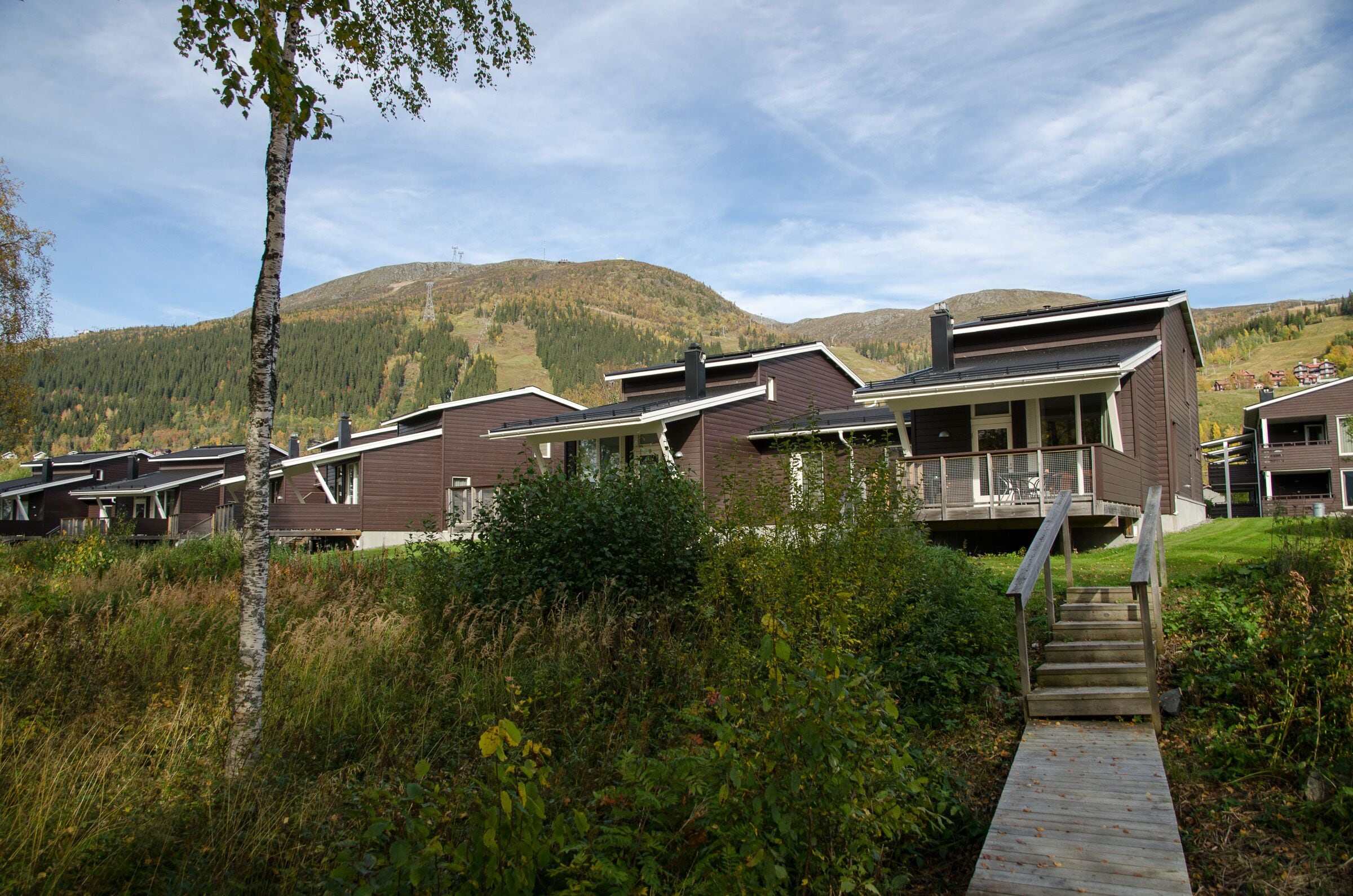 A row of modern cabins with a wooden deck leading to a covered porch, set against a backdrop of hills and a clear sky.